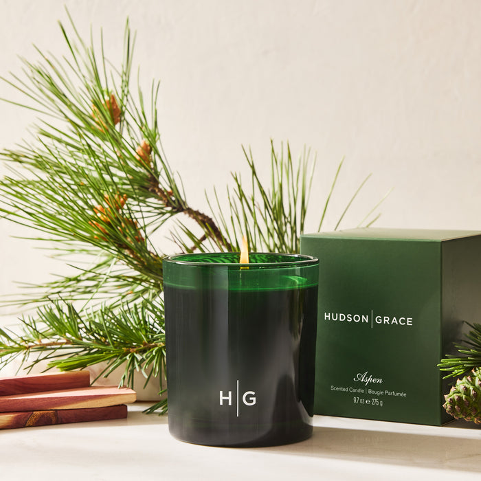 Hudson Grace Aspen Scented Candle in a dark green glass vessel with a matching box, surrounded by pine branches.