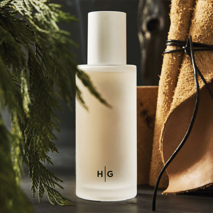 Hudson Grace Bleecker Scented 3-Wick Candle in a frosted glass vessel with HG logo, surrounded by greenery and leather.