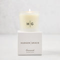 Hudson Grace Greenwich Scented Votive, a lit candle in a frosted glass vessel with "HG" logo, on its white box.