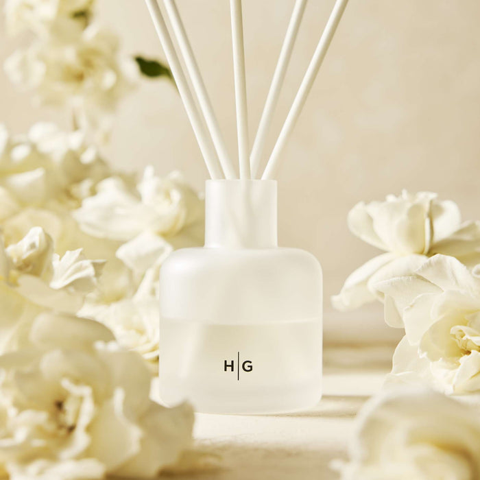 Hudson Grace Greenwich reed diffuser, frosted white glass bottle with HG logo, surrounded by white gardenia flowers.