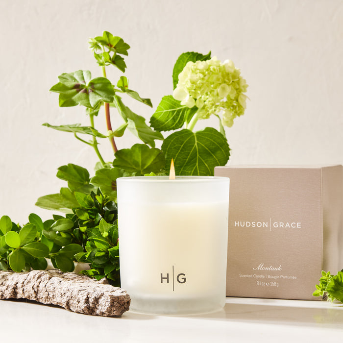 Hudson Grace Montauk scented candle in a frosted glass jar with a beige box, green plants, and wood.