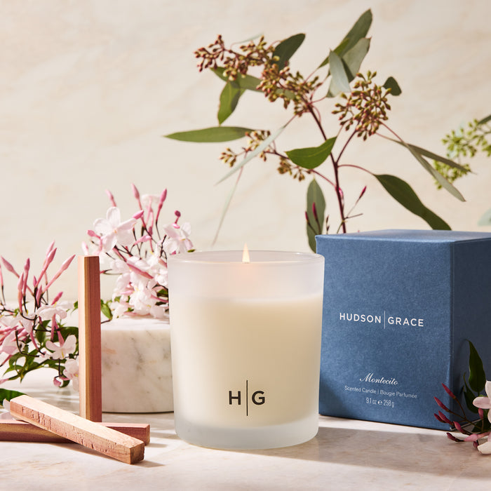Hudson Grace Montecito 3-wick candle in a frosted glass vessel, lit, with blue box, wood sticks, and flowers.