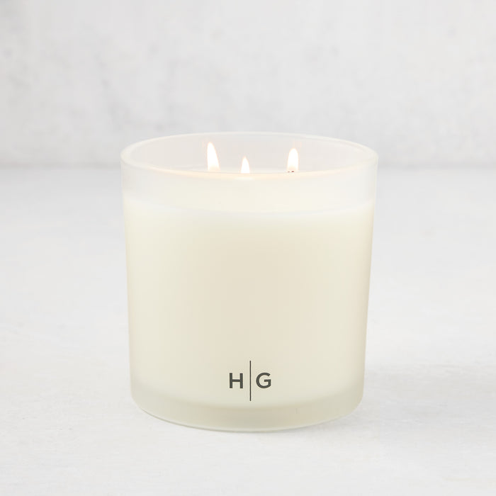 Hudson Grace Montecito 3-wick candle in a frosted glass vessel with "HG" logo, lit on a white surface.