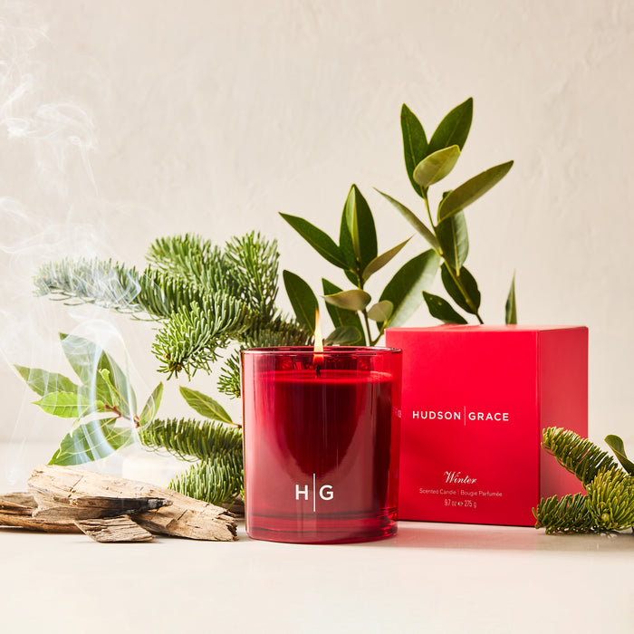 Hudson Grace Winter Scented Candle in a red glass vessel with a red box, surrounded by evergreens and wood.