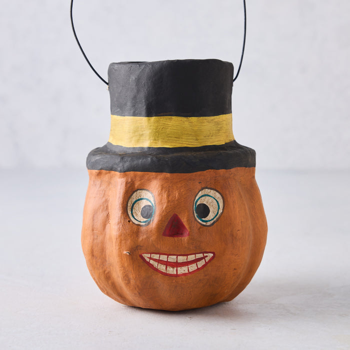 Vintage-style papier-mâché pumpkin candy bucket wearing a black top hat with a yellow band and wire handle.