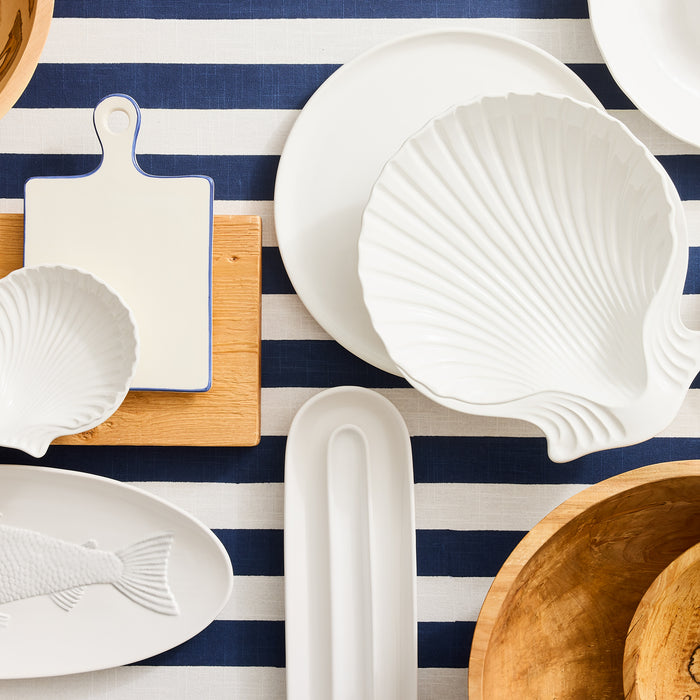 White ceramic olive serving tray with a shell design, 16" long, on a blue and white striped tablecloth.
